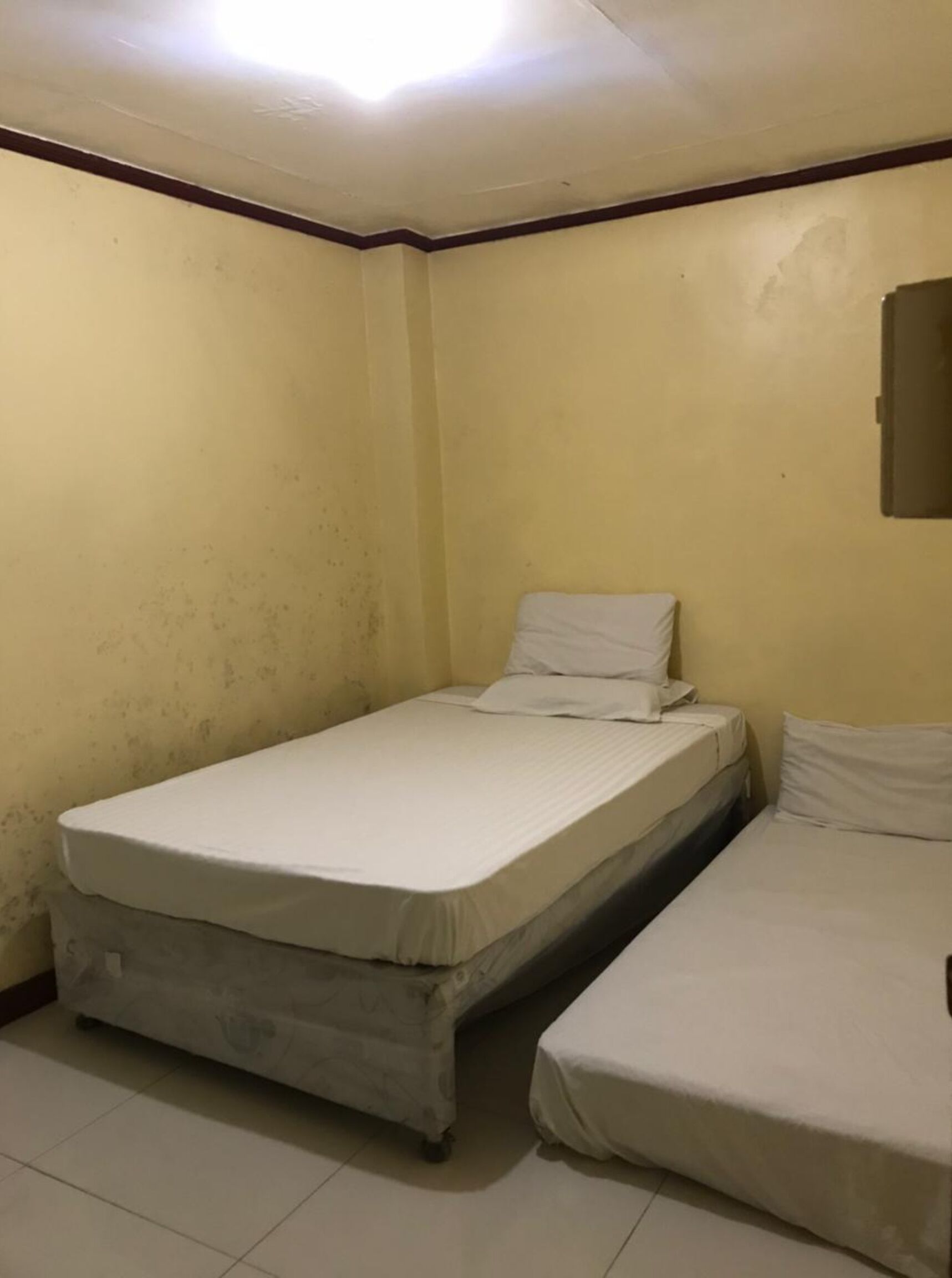 Ein kleines Zimmer mit zwei Betten, schlichten Wänden und einer Deckenleuchte.