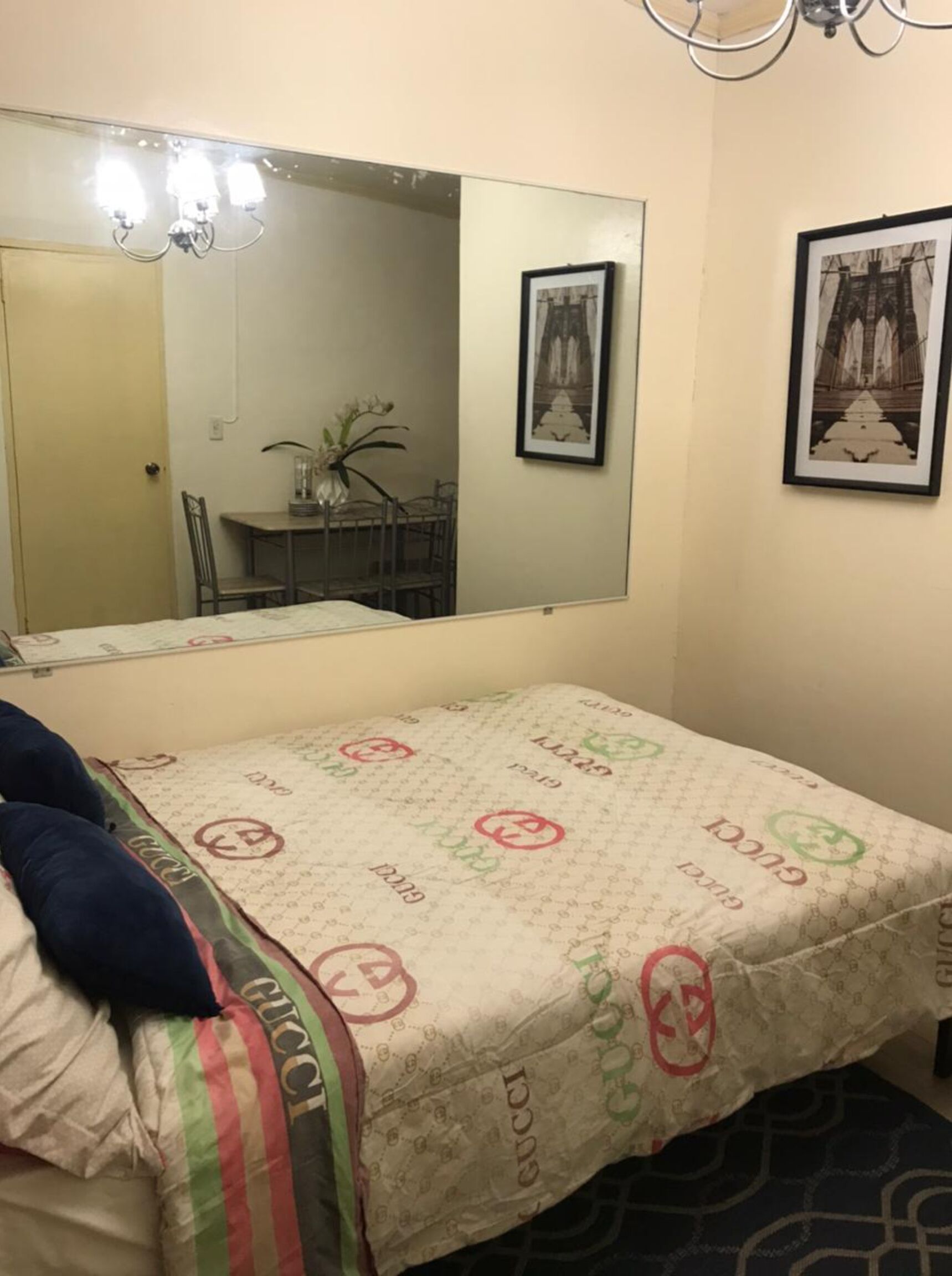 Un dormitorio con un espejo grande, una cama con edredón estampado de Gucci, una silla, una mesa y dos cuadros en la pared.