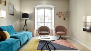 Uma sala de estar com um sofá azul, duas poltronas, uma mesa de centro redonda, um abajur e uma janela grande com vista.