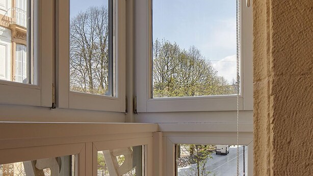 Quarto com vista para a rua através de grandes janelas, uma mesa com um livro e uma porta com puxador.