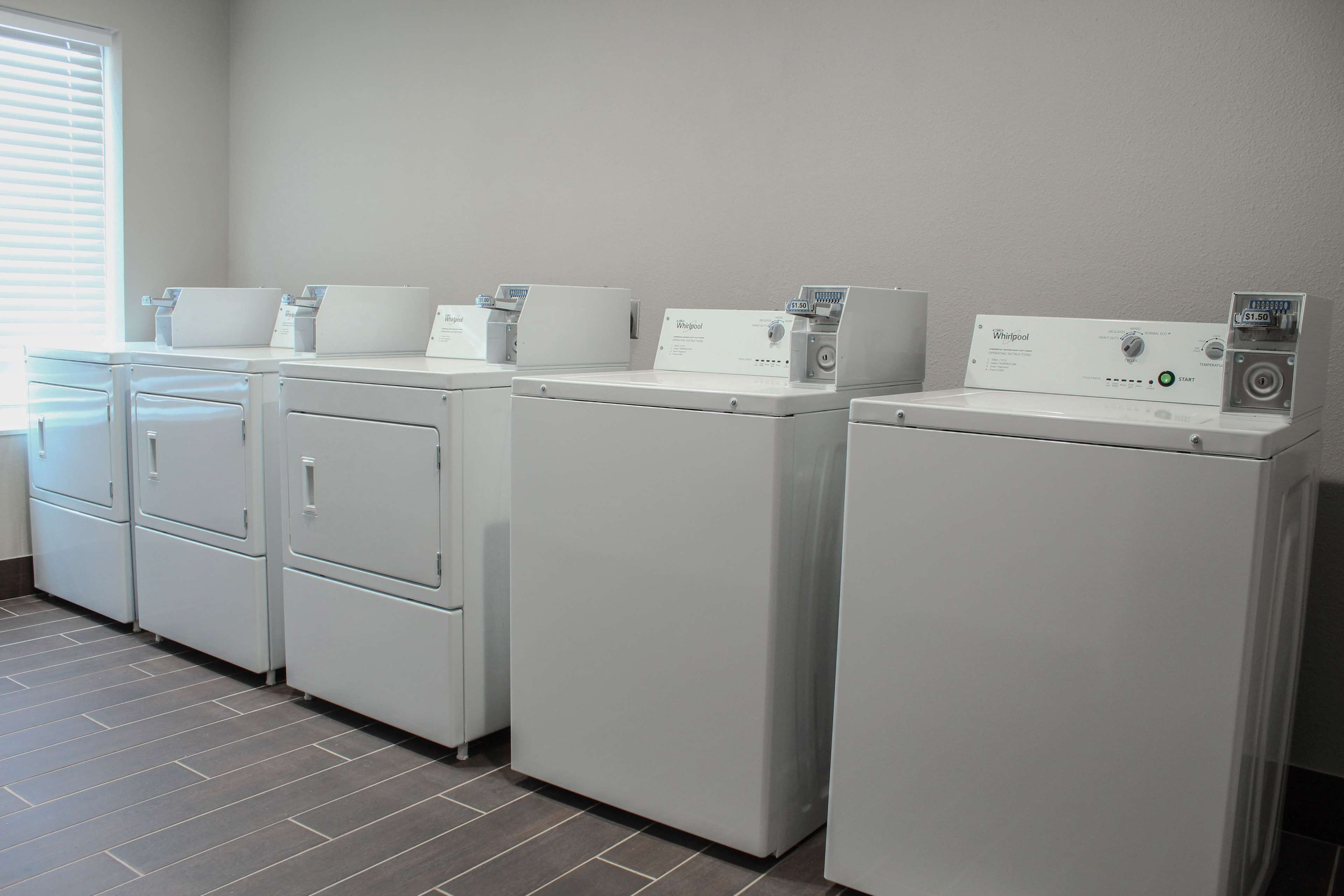 A row of white Whirlpool washers in a laundry room with a tiled floor and a window with blinds.