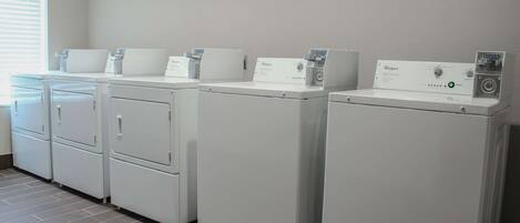A row of white Whirlpool washers in a laundry room with a tiled floor and a window with blinds.