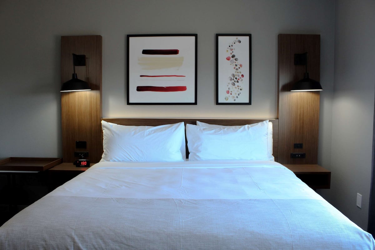 Quarto de hotel com uma cama grande, duas mesas de cabeceira, um criado-mudo e duas luminárias fixadas na parede.