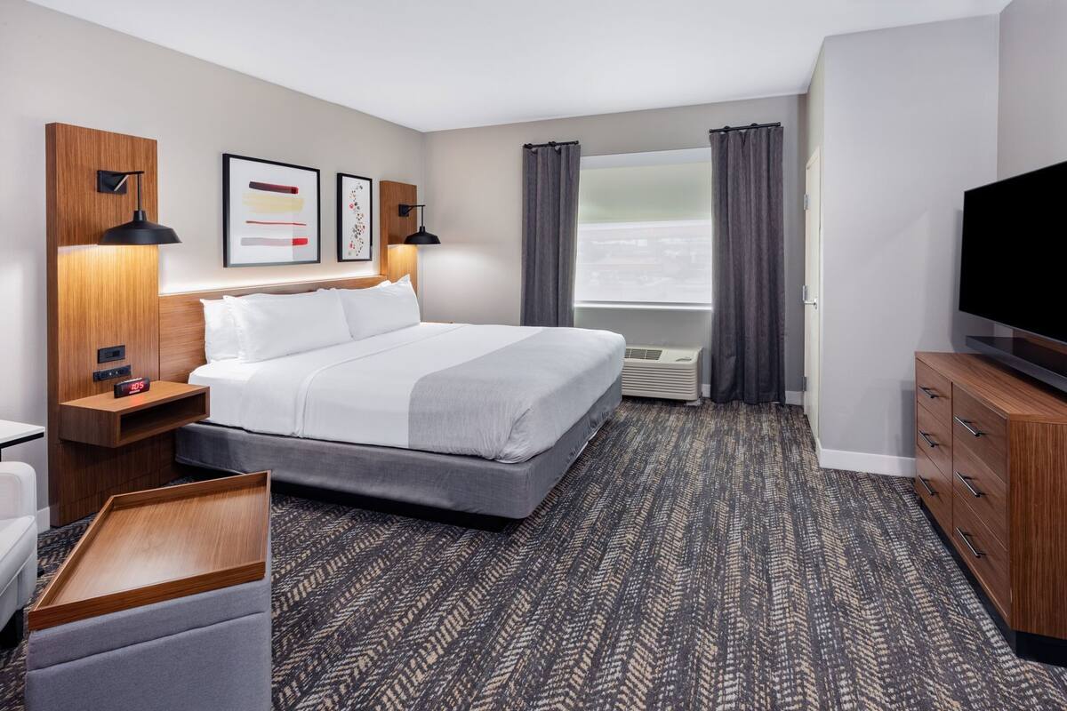 Quarto de hotel com uma cama, televisão, escrivaninha, cadeira, mesa de cabeceira, relógio e janela com cortinas.