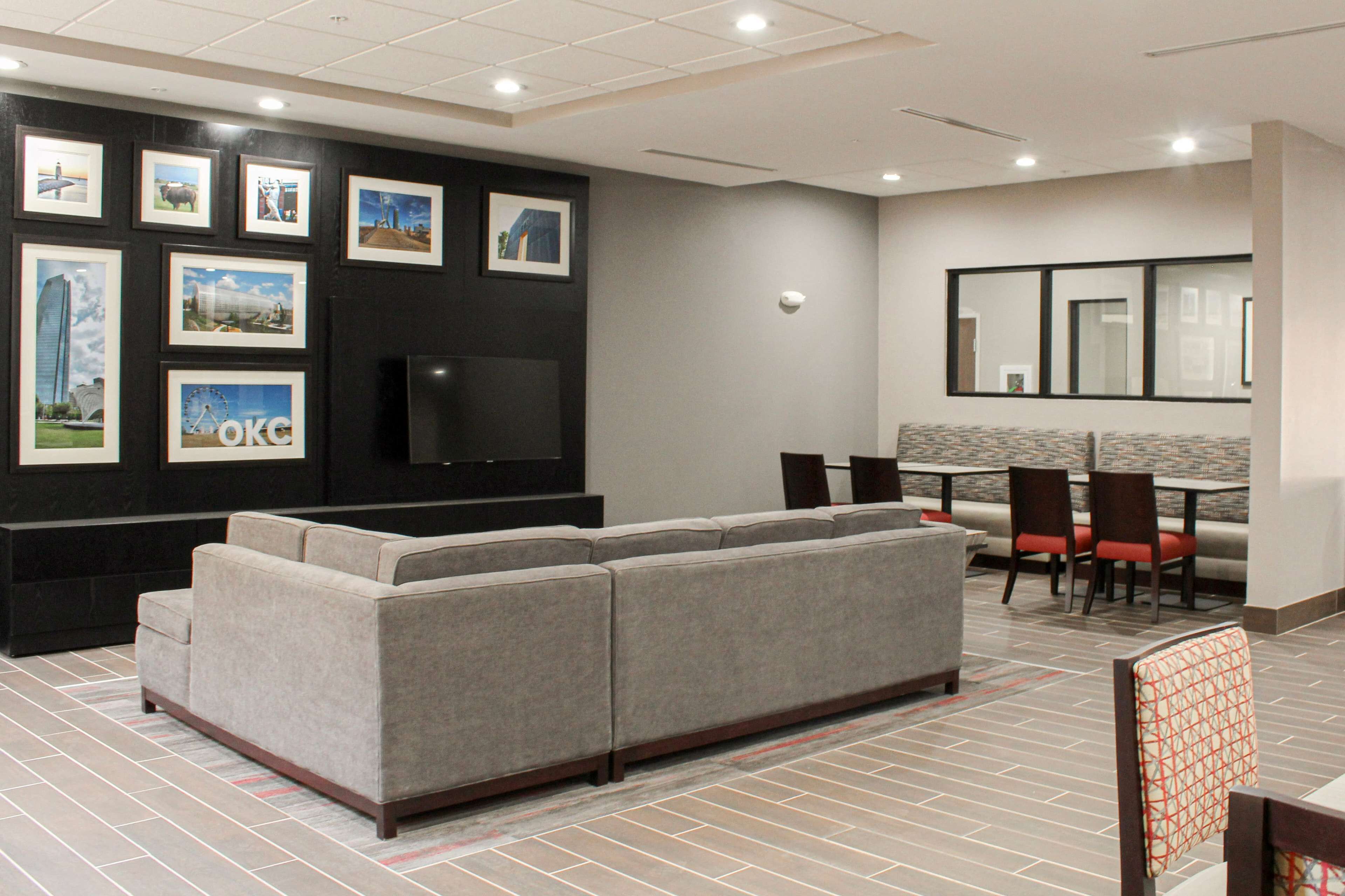 A modern hotel lobby with a large gray sofa, a flat-screen TV, and a dining area with red chairs.