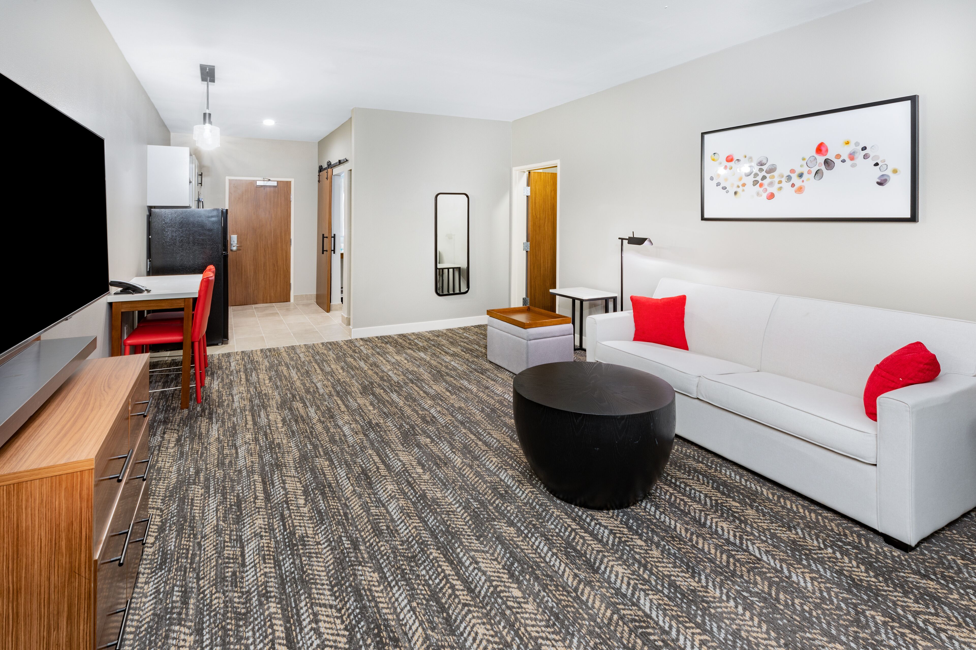 A modern living room with a flat-screen TV, a wooden desk, a red chair, a white sofa with red cushions, a black coffee table, and a patterned carpet.