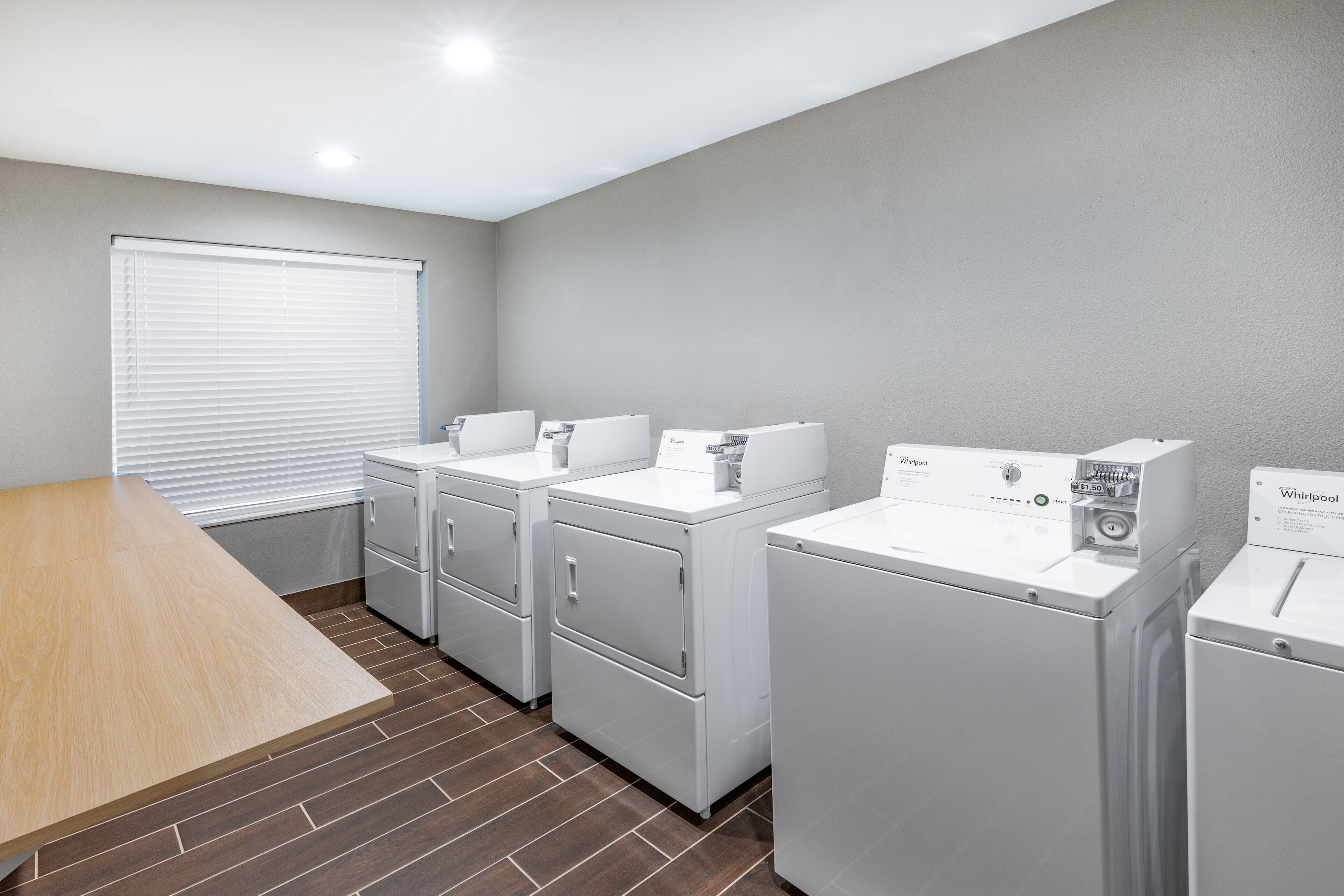 Una sala de lavandería con cuatro lavadoras y secadoras Whirlpool, una mesa de madera y una ventana con persianas.