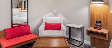 A hotel room with a bed, a white armchair with a red pillow, a red sofa, a small wooden table, and a wall-mounted nightstand with a clock.