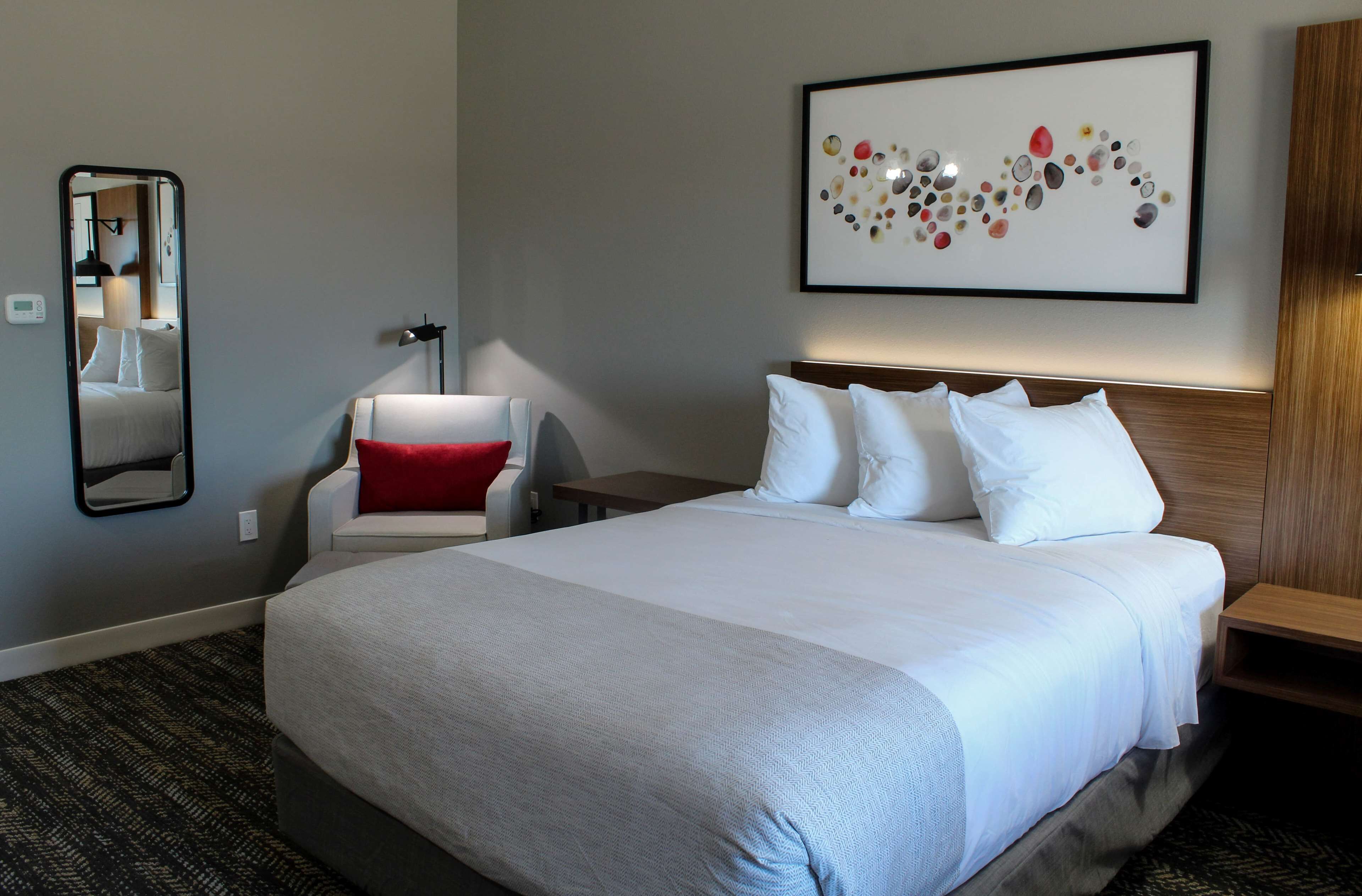 A hotel room with a large bed, a bedside table, a chair with a red cushion, a wall-mounted mirror, and a framed artwork above the bed.