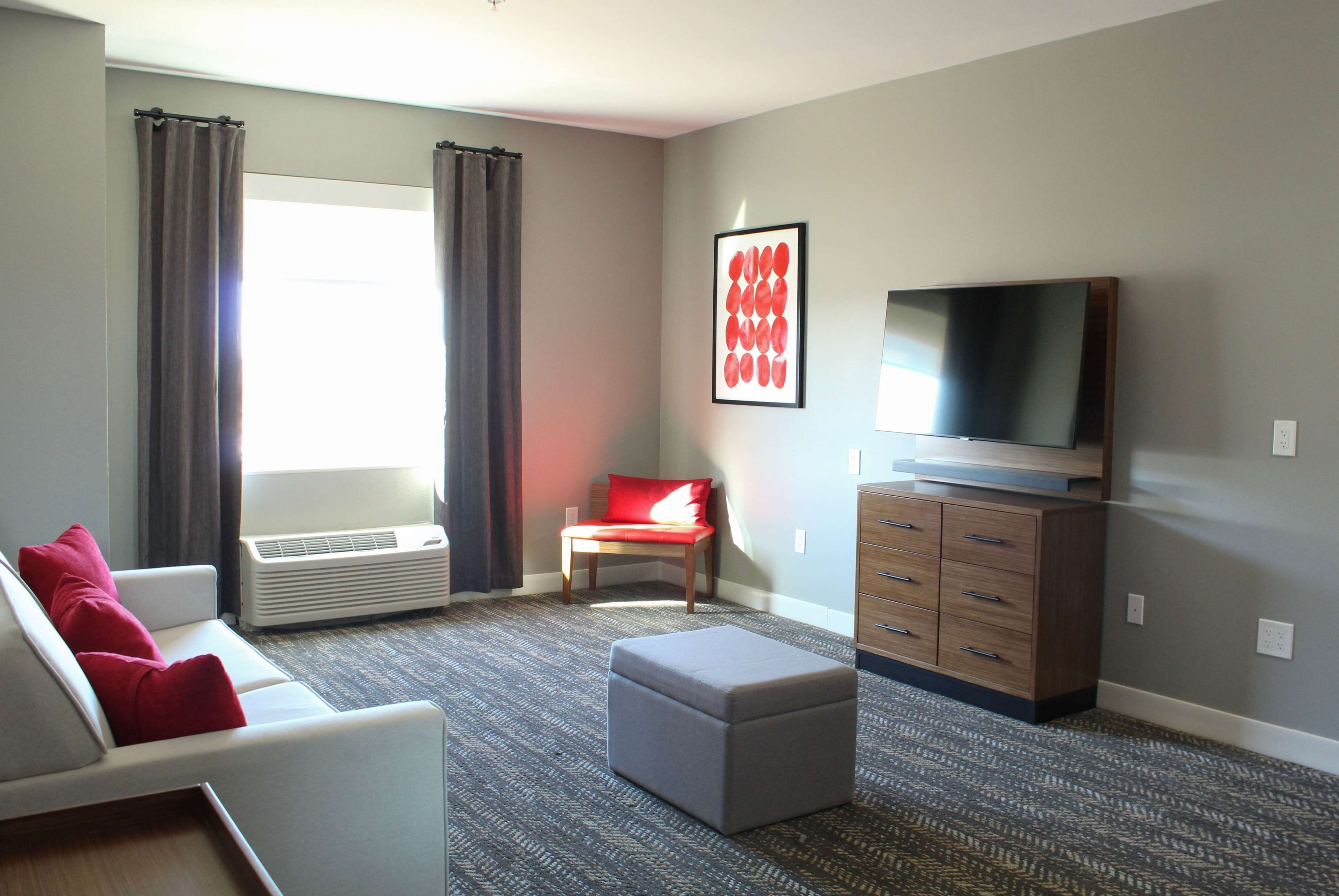 Un salon moderne avec un canapé gris, des coussins rouges, un pouf gris, un téléviseur à écran plat, un meuble en bois et une grande fenêtre avec des rideaux.