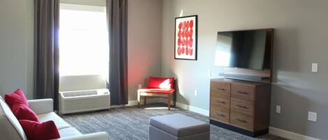 Un salon moderne avec un canapé gris, des coussins rouges, un pouf gris, un téléviseur à écran plat, un meuble en bois et une grande fenêtre avec des rideaux.