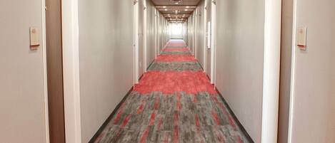 Un pasillo de hotel con alfombra roja y baldosas grises, que conduce a una habitación luminosa al final.