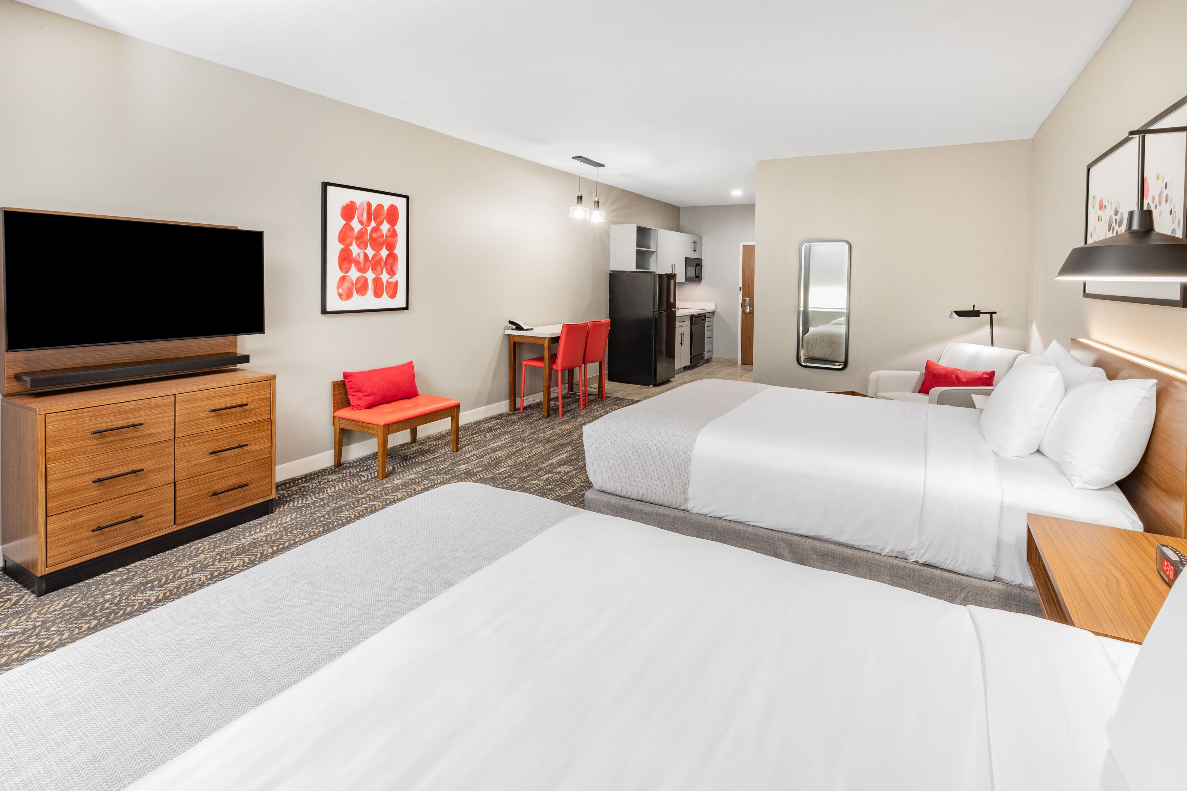 A hotel room with two beds, a TV, a desk with chairs, a kitchen area, and a mirror.