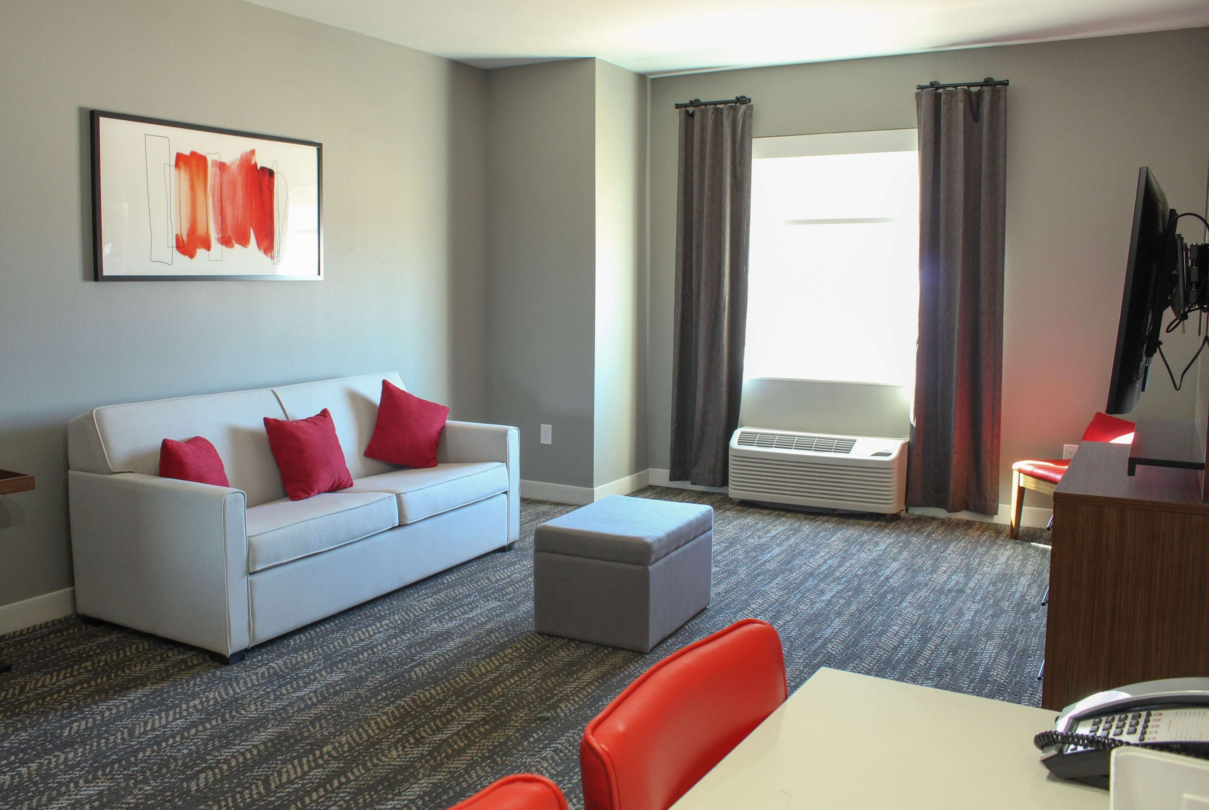 Une chambre d’hôtel moderne avec un canapé gris, des coussins rouges, un pouf gris, un bureau blanc et un téléviseur à écran plat.