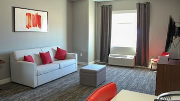 Une chambre d’hôtel moderne avec un canapé gris, des coussins rouges, un pouf gris, un bureau blanc et un téléviseur à écran plat.