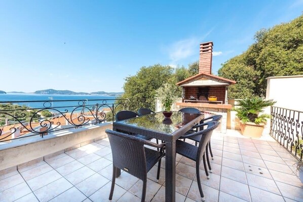Una terraza con una mesa de vidrio y sillas de mimbre, con vistas panorámicas al mar y las colinas.