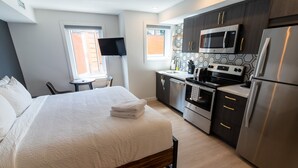 Habitación de hotel moderna con una cama grande, un escritorio pequeño, televisor de pantalla plana, cocina con electrodomésticos de acero inoxidable y ventana con vistas al exterior.