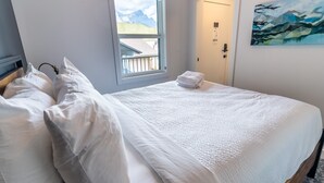Een net opgemaakt bed met witte lakens en een gevouwen handdoek, een raam met uitzicht op bergen, en een schilderij aan de muur.