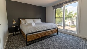 A bedroom with a large bed, a nightstand, a lamp, and a view of the outdoors through a sliding glass door. - Basecamp Suites Canmore I Two Bedroom Apartment (Canmore)