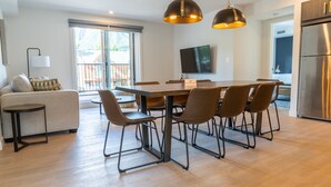 A modern kitchen with a stainless steel refrigerator, a dining area with a wooden table and chairs, and a sofa with a patterned throw. - Basecamp Suites Canmore I Two Bedroom Apartment (Canmore)