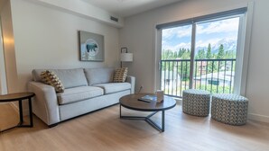 A modern living room with a sofa, coffee table, and ottomans, a large window with a mountain view, and a framed artwork on the wall. - Basecamp Suites Canmore I Two Bedroom Apartment (Canmore)