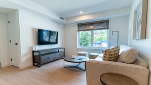 Ein modernes Wohnzimmer mit Flachbildfernseher, einer Couch, einem Couchtisch und einem Fenster mit Jalousien.