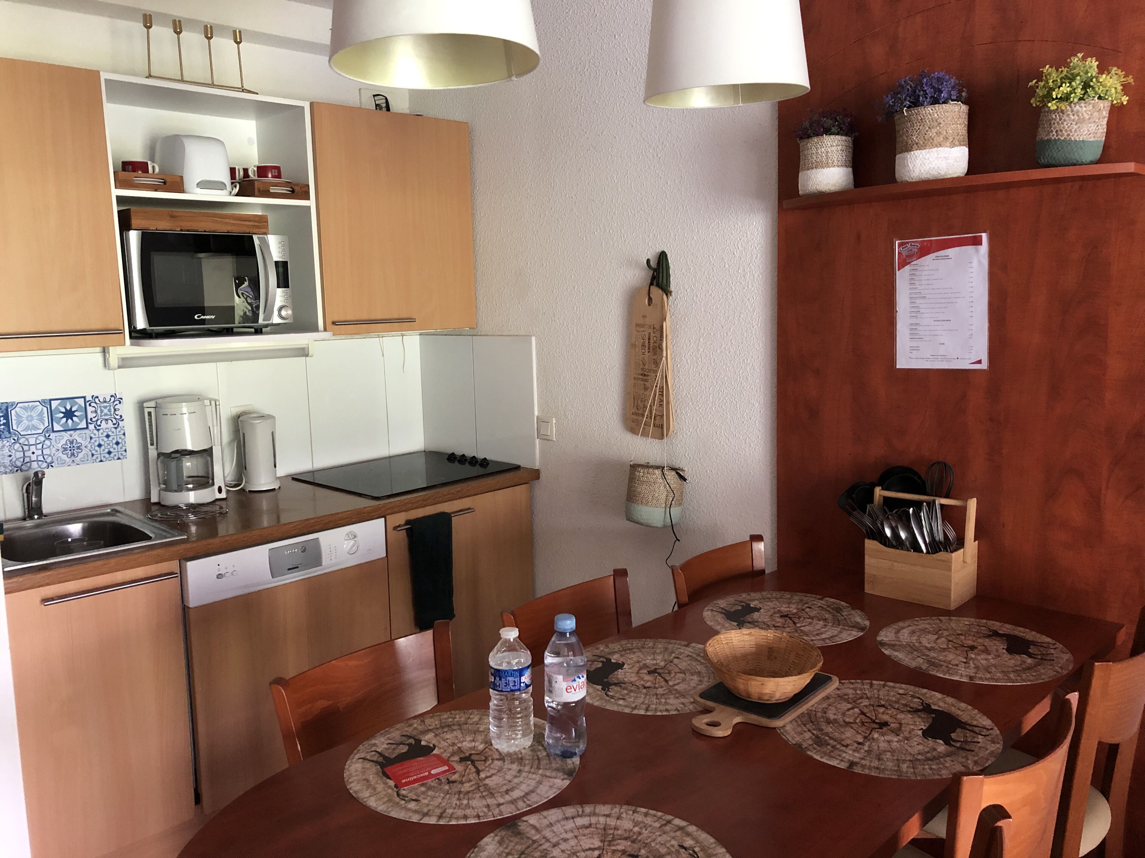 A compact kitchen with a microwave, coffee maker, and a sink. There are wooden cabinets, a dining table with placemats, and a few potted plants on a shelf.