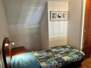 Una habitación pequeña con una cama individual, cabecera de madera, lámpara de noche e imagen en un marco en la pared.