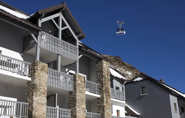 Un edificio de varios pisos con balcones, pilares de piedra y un teleférico al fondo.