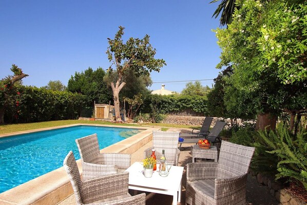 A swimming pool with wicker chairs and a table, surrounded by greenery and a stone wall.