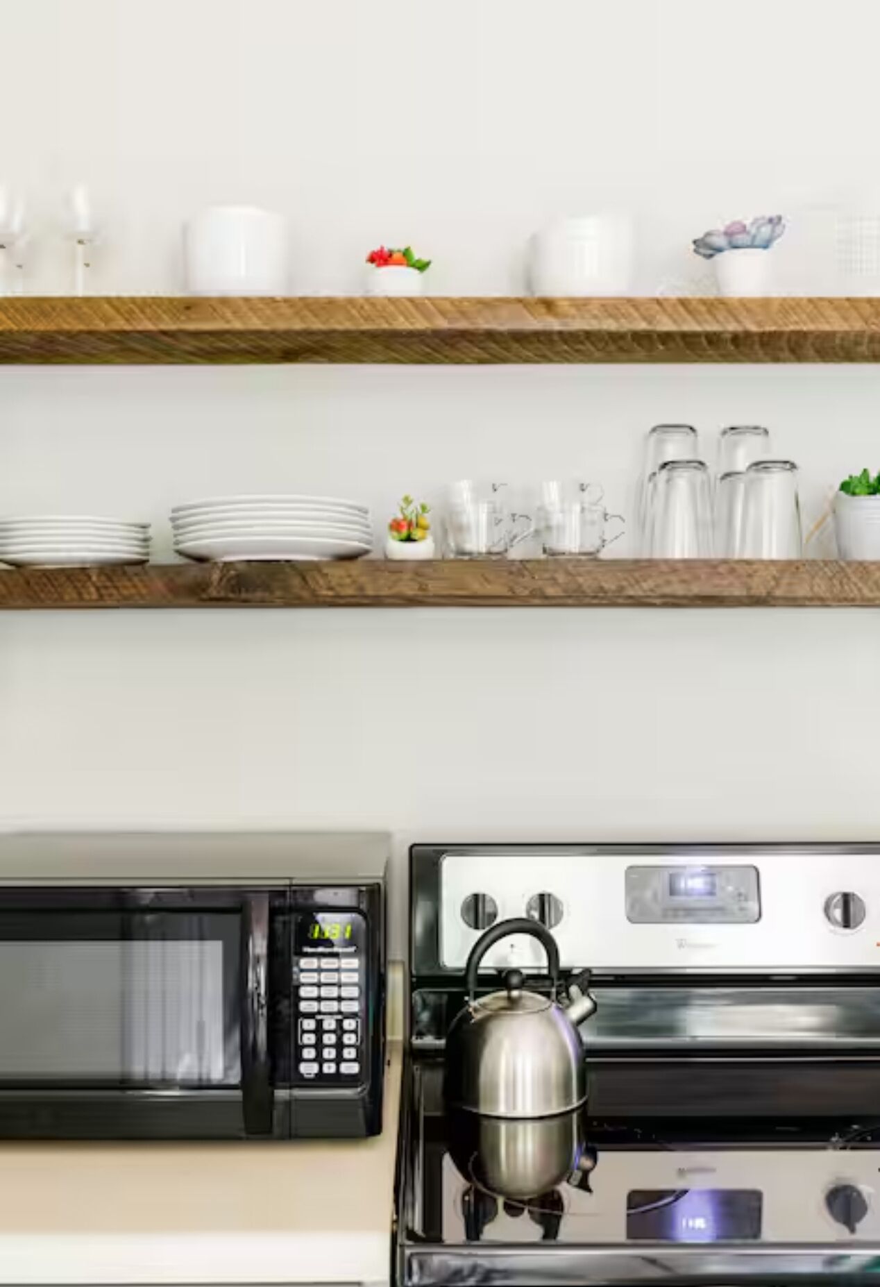 Une cuisine équipée d’un four à micro-ondes, d’une bouilloire et d’une cuisinière. Des assiettes et des tasses blanches sont rangées sur des étagères au-dessus de la cuisinière.