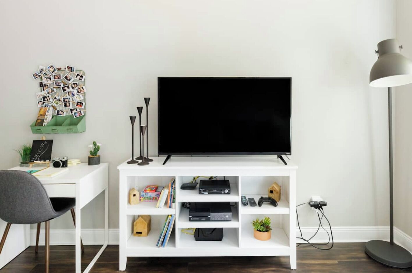Una sala moderna con una televisión de pantalla plana sobre un mueble blanco, un escritorio con una silla, una lámpara de pie y un collage de fotos colgado en la pared.