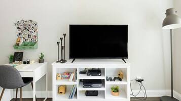 Una sala moderna con una televisión de pantalla plana sobre un mueble blanco, un escritorio con una silla, una lámpara de pie y un collage de fotos colgado en la pared.