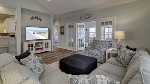 A spacious living room with a television displaying an island getaway rental ad, a dining area with a wooden table and chairs, and a cozy seating area with patterned cushions.