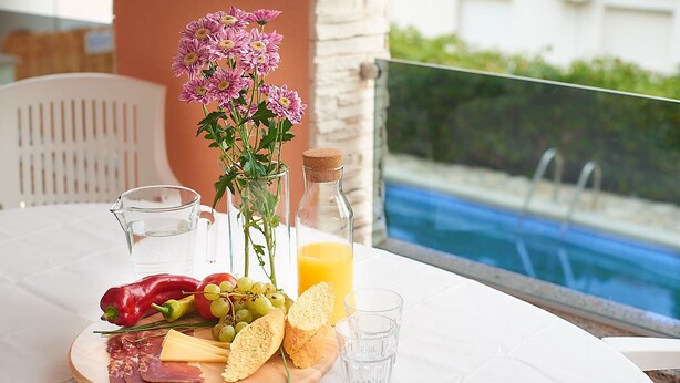 A table with a cheese board, fruits, bread, and a bottle of orange juice, set against a poolside view. - Villa Adria II, apartment B3, with 2 swimming pools (Pag)