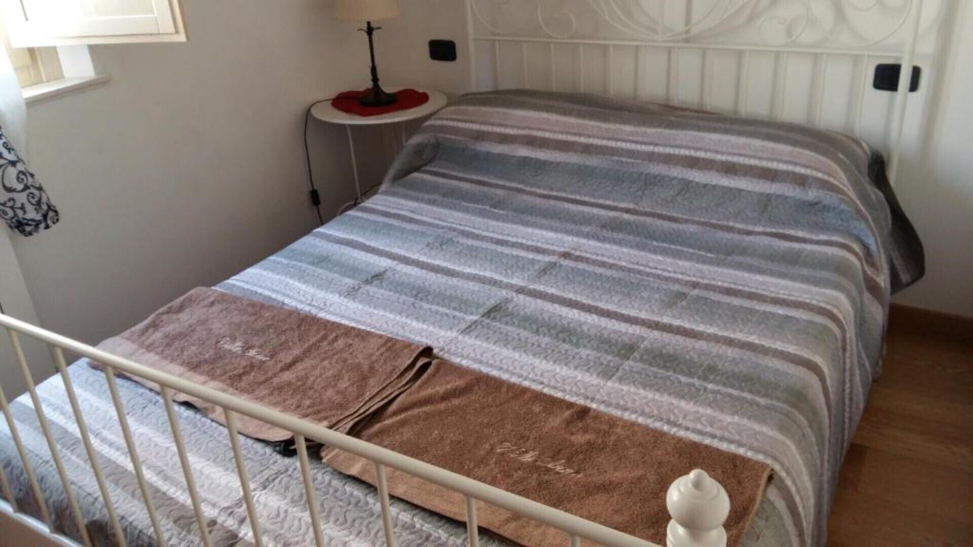 Un lit avec une couverture à rayures et un plaid marron, une table de chevet avec une lampe et une tête de lit blanche.