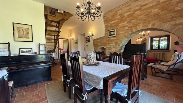 Un comedor con una mesa dispuesta para cuatro personas, sillas de madera, una araña, un piano y una pared de piedra.