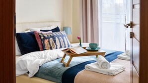 Ein ordentlich bezogenes Bett mit einer blauen und weißen Bettdecke, ein Nachttisch aus Holz mit einem Buch, einer Tasse und einer Tulpe.
