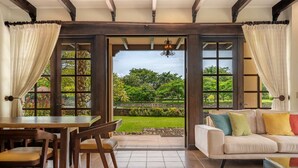 A spacious room with a dining area, a sofa, and a view of a lush garden through large windows.