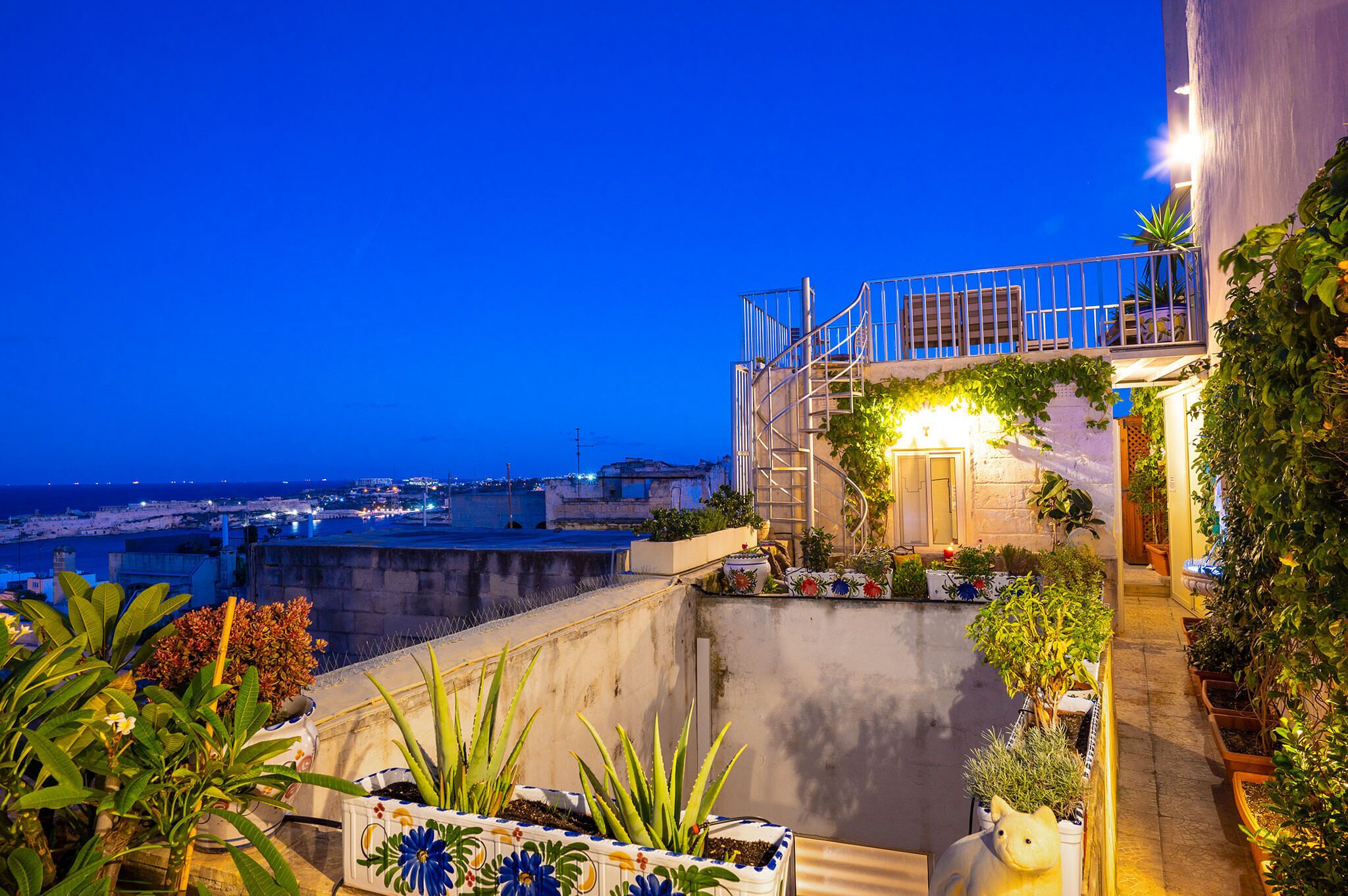 Una terrazza sul tetto con piante in vaso, una scala e vista sulla città al crepuscolo.
