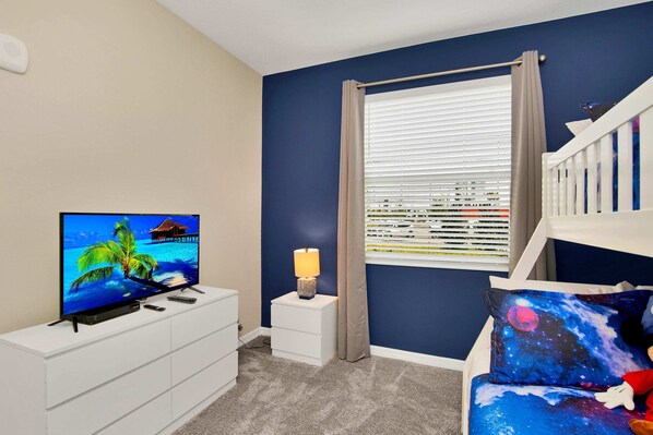 A bedroom with a TV on a white dresser, a nightstand with a lamp, a bed with space-themed bedding, and a window with blinds.