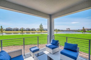 A covered outdoor seating area with wicker furniture and blue cushions, overlooking a green lawn and a body of water.