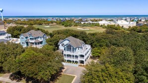 A large, two-story house with a balcony, surrounded by dense trees and a golf course. - Seadog:  Private Pool, Hot Tub, Rec Room (Corolla)