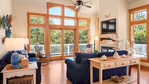 A spacious living room with a high ceiling, large windows, a ceiling fan, a television, and a fireplace. - Seadog:  Private Pool, Hot Tub, Rec Room (Corolla)