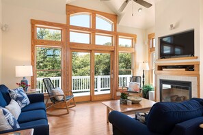 A spacious living room with a large wooden-framed window, a fireplace, and a television. - Seadog: Private Pool, Hot Tub, Rec Room (Corolla)