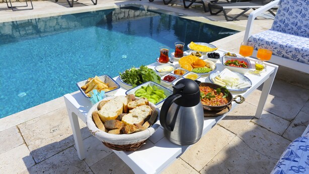 Una mesa junto a la piscina con diversos alimentos y bebidas, rodeada de sillones de descanso.