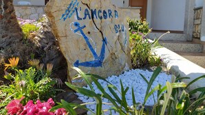En stenskilte med '#LANCORA' og et ankersymbol, omgivet af planter og hvide sten.