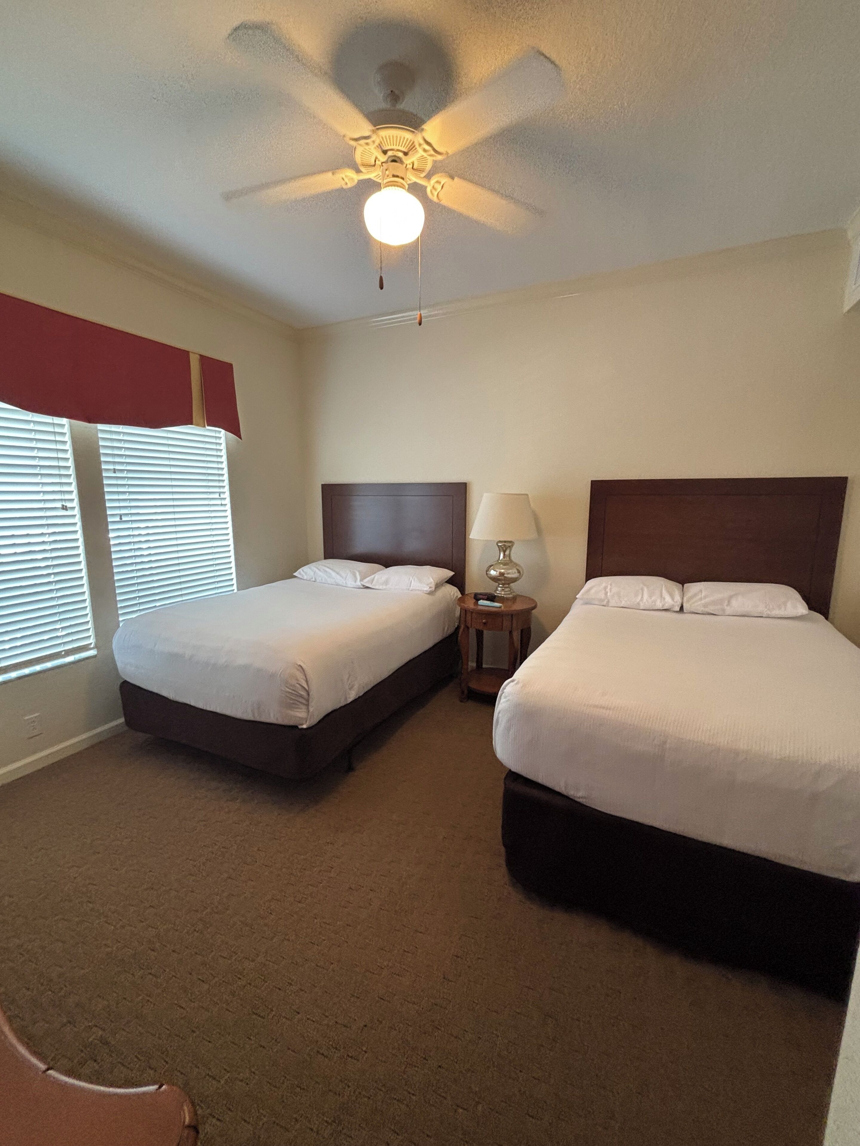 A hotel room with two beds, a ceiling fan, a bedside table with a lamp, and a window with blinds.