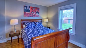 Une chambre à coucher avec un lit en bois, deux lampes de chevet, un ventilateur de plafond, une fenêtre donnant sur un beau paysage et un tableau coloré accroché au mur.