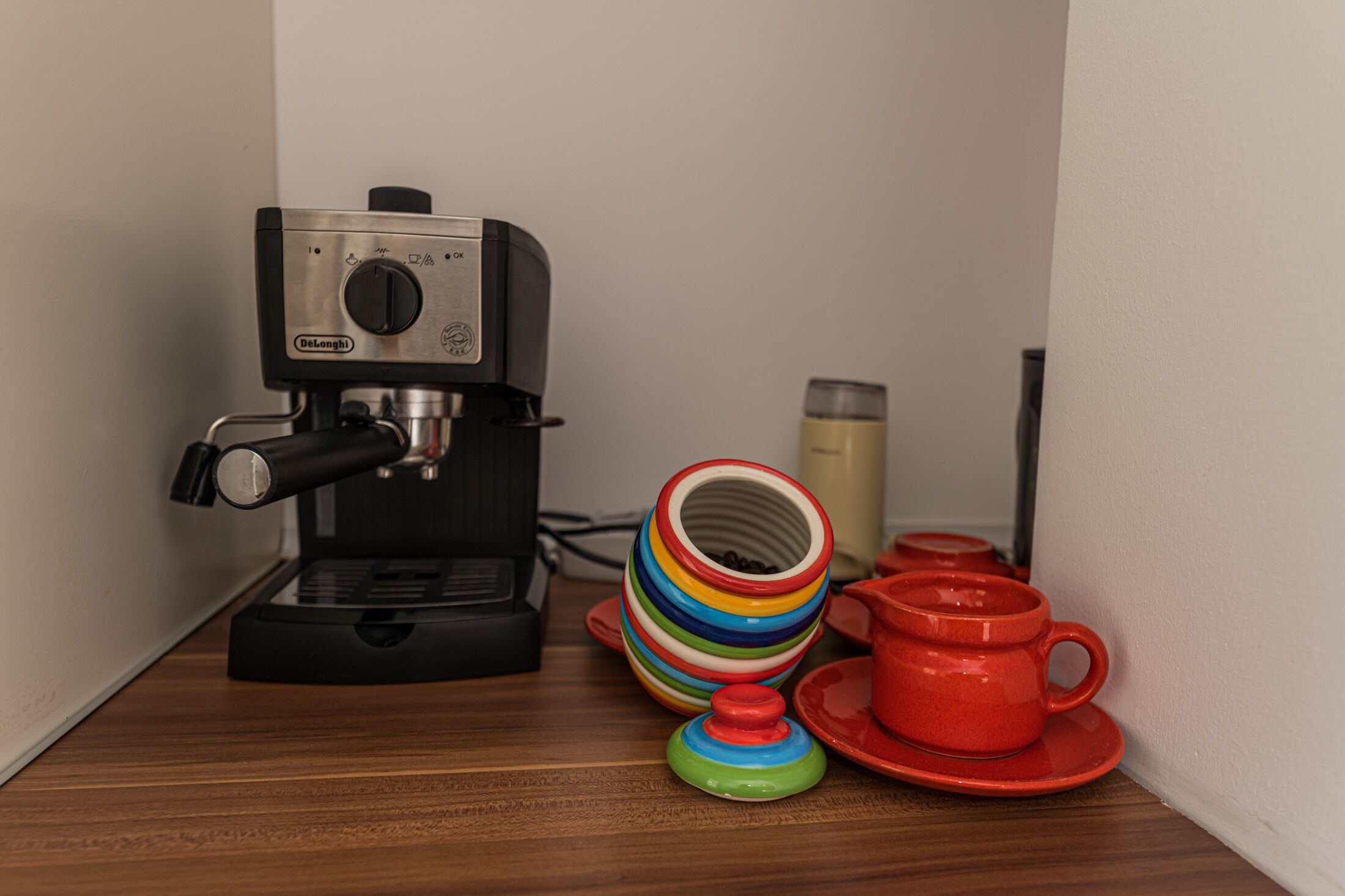 A DeLonghi espresso machine on a wooden surface with a colorful stack of cups and a thermos.